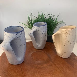 May include: Three ceramic mugs with distinctive handles and speckled patterns. Two are light blue with dark blue speckles and rims, the third is beige with brown speckles and a brown rim. They are on a large, round, wooden tray.