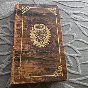 May include: An antique brown leather-bound book with gold-coloured filigree borders. The front cover features a central emblem with a shield, laurel leaves, and the text 1845 PENSIONNAT MAQUART A REIMS embossed in gold lettering on the weathered surface.