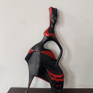 May include: A stylized wall sculpture of a woman in a long dress. The sculpture is primarily black with red accents on the head, bodice, and skirt. The figure has a long, elegant neck and a flowing skirt, creating a graceful silhouette.