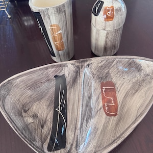 Peut inclure: Un ensemble en céramique composé d'un grand gobelet cylindrique, d'un récipient en forme de bouteille avec bouchon et d'un plat de service triangulaire divisé. Chaque pièce présente une finition beige et grise avec des accents peints.