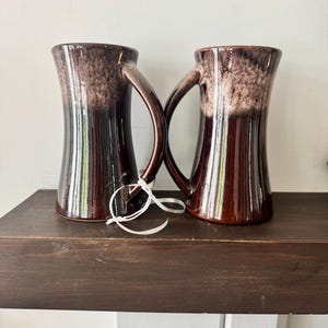 Può includere: Due tazze in ceramica alte e svasate con una smaltatura marrone scuro lucida alla base che sfuma in una finitura testurizzata screziata color marrone chiaro e crema sulla parte superiore. Ogni tazza ha un manico curvo e un cordino bianco.