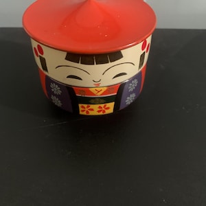Könnte beinhalten: Ein roter und weißer Keramikbehälter mit einem konischen Deckel. Der Behälter zeigt ein gemaltes Bild einer Frau in einem traditionellen japanischen Kimono mit einem Blumenmuster. Die Frau hat schwarzes Haar und ein lächelndes Gesicht.