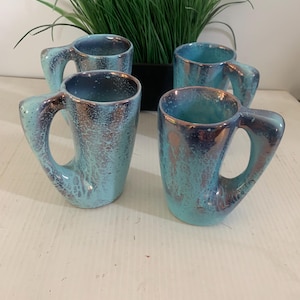 May include: Four ceramic mugs with a distinctive handle design. The mugs feature a turquoise and dark blue glaze with a textured finish. The mugs are grouped together, with a green plant visible in the background.