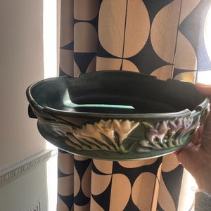 Puede incluir: Un cuenco redondo de cerámica verde oscuro con un relieve moldeado de flores de tonos claros alrededor del borde exterior. El cuenco tiene un acabado liso y brillante y una parte superior ancha y abierta con un diseño de borde orgánico irregular.