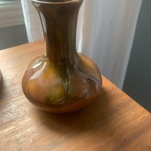 May include: A small, glazed ceramic vase with a bulbous base and a narrow neck. The vase has a glossy finish with a blend of brown, green, and gold tones. It sits on a wooden surface, showcasing its unique shape and color.