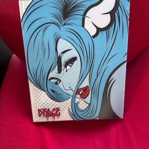 May include: A rectangular book featuring a pop art illustration of a person with long blue hair, dark eyelashes, and red lips. The background has a light blue polka dot pattern. The text D*FACE is printed in red graffiti-style lettering at the bottom.