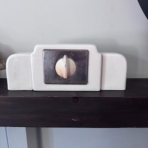 Op de afbeelding: Een rechthoekig wit keramisch object met afgeronde randen en een getrapt bovenprofiel. Centraal op de voorzijde bevindt zich een vierkante metalen plaat met een ronde draaiknop met verticale ribbels in een lichte metaalkleur.