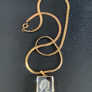 Peut inclure: Collier doré avec un pendentif rectangulaire représentant un camée du profil d'une femme. Le pendentif est encadré d'or. La chaîne est de conception plate et tissée. Le collier est présenté sur une surface sombre.