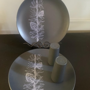 May include: Two grey ceramic plates and two matching salt and pepper shakers. Each plate features a white pine branch design. The plates are displayed on a metal stand. The salt and pepper shakers are cylindrical.