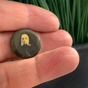 May include: A circular pin with a dark background, displaying a portrait of a woman with a yellow veil and black clothing. A small cross is visible. The pin is held in a hand, with a blurred green backdrop.