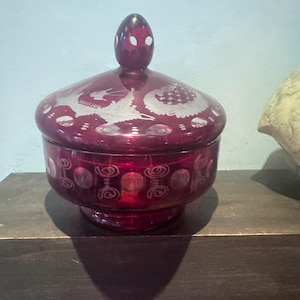 Peut inclure: Un bonbonnier en verre rouge profond avec un couvercle assorti doté d'un bouton pointu. La surface extérieure est ornée de motifs blancs gravés, incluant un dessin d'oiseau sur le couvercle et des cercles et spirales autour de la base.
