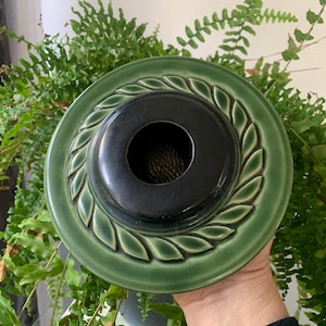 May include: A green ceramic lid with a black center and a decorative leaf design around the edge. The lid has a hole in the center.