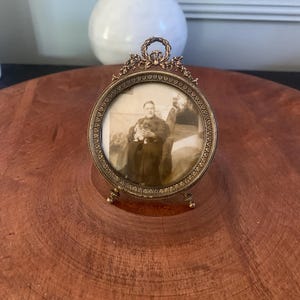 Peut inclure: Un cadre photo ovale antique de couleur bronze avec un motif floral et des volutes détaillés. Le cadre contient une photographie en noir et blanc d'une personne tenant un petit chien. Le cadre a un petit support et une boucle décorative en haut.