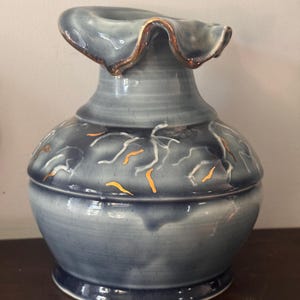 May include: A ceramic vase with a ruffled, flared rim featuring a metallic gold edge. The body is a gradient of slate blue and grey, decorated with scattered, abstract white and metallic gold brushstroke patterns across the rounded midsection.
