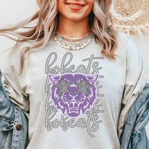 May include: A light gray sweatshirt with the word "bobcats" in a script font. A purple bobcat graphic with glitter star-shaped sunglasses is centered on the front. The word "bobcats" is repeated in a vertical pattern behind the bobcat.