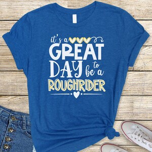May include: A royal blue t-shirt with a white graphic that says "it's a great day to be a Roughrider" with yellow hearts and a yellow outline.