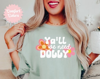 May include: A light green t-shirt with the text "Ya'll we need Dolly" in white with a pink and orange flower graphic on either side of the text.