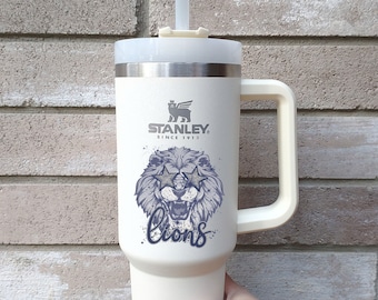 May include: White stainless steel travel mug with a black and white lion graphic wearing sunglasses and the text "lions". The mug has a black handle and a black lid with a straw.