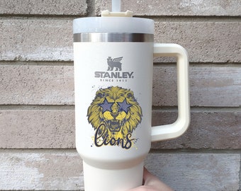 May include: White Stanley travel mug with a lion wearing sunglasses and the text "lions" printed on the side. The mug has a black handle and a straw.