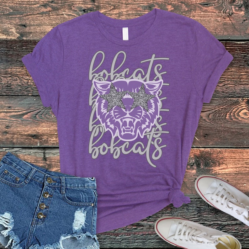 May include: Purple t-shirt with a graphic of a bobcat wearing star-shaped sunglasses. The word "bobcats" is written above and below the graphic in a cursive font. Denim shorts and white sneakers are visible.