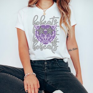 May include: White t-shirt featuring a purple bobcat graphic wearing star-shaped sunglasses. The word "bobcats" is repeated in a script font above and below the graphic. The shirt is a crew neck style.