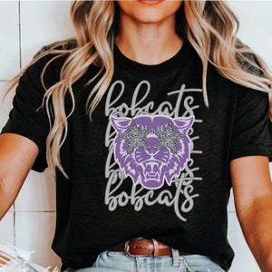 May include: Dark gray t-shirt featuring a purple bobcat graphic with glitter star-shaped glasses. The word "bobcats" is written in a cursive font above and below the graphic. The shirt has a crew neck and short sleeves.