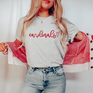May include: A gray t-shirt with the word "cardinals" written in red glitter lettering with a red glitter heart next to it. The shirt is being worn by a person with long blonde hair and a red cardigan.