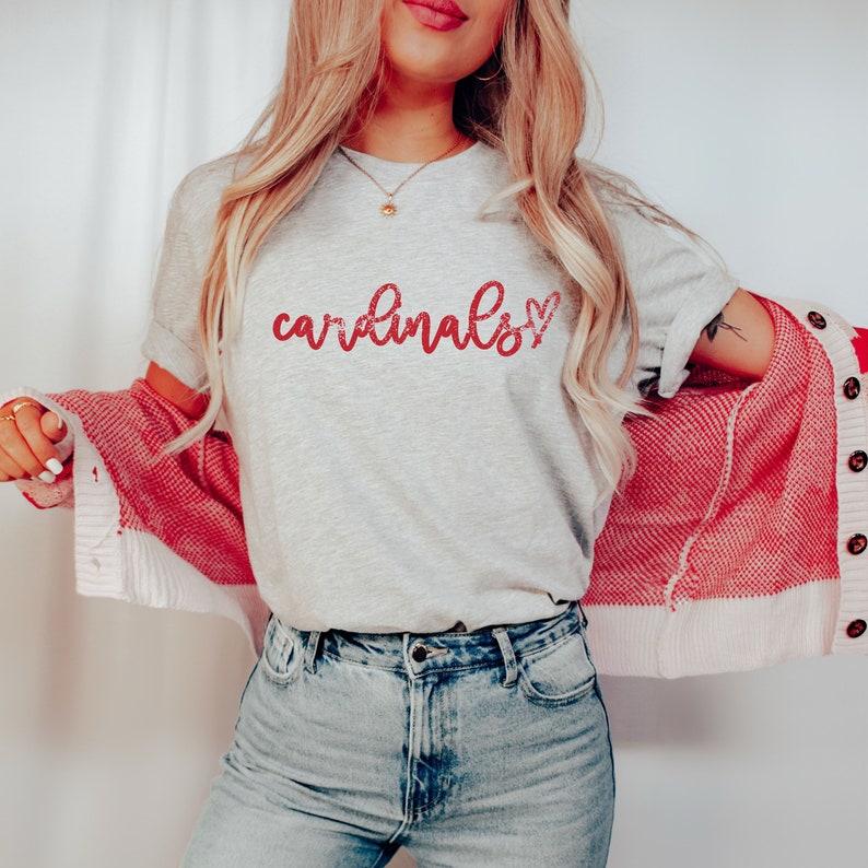 May include: A gray t-shirt with the word "cardinals" written in red glitter lettering with a red glitter heart next to it. The shirt is being worn by a person with long blonde hair and a red cardigan.