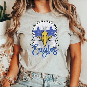 Pu&ograve; includere: Una maglietta grigia con una grafica di un'aquila blu e bianca usurata e la parola "Eagles" in lettere blu.