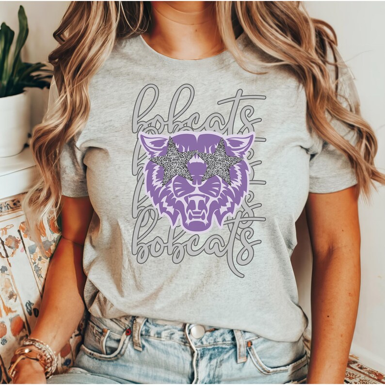 May include: A light gray t-shirt featuring a purple bobcat graphic with star-shaped sunglasses. The word "bobcats" is repeated in a script font. The shirt is a crew neck style.