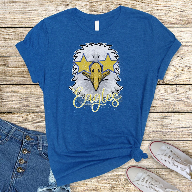 Pu&ograve; includere: Una t-shirt blu reale con una grafica usurata di un'aquila che indossa occhiali da sole e la parola "Eagles" in lettere gialle.
