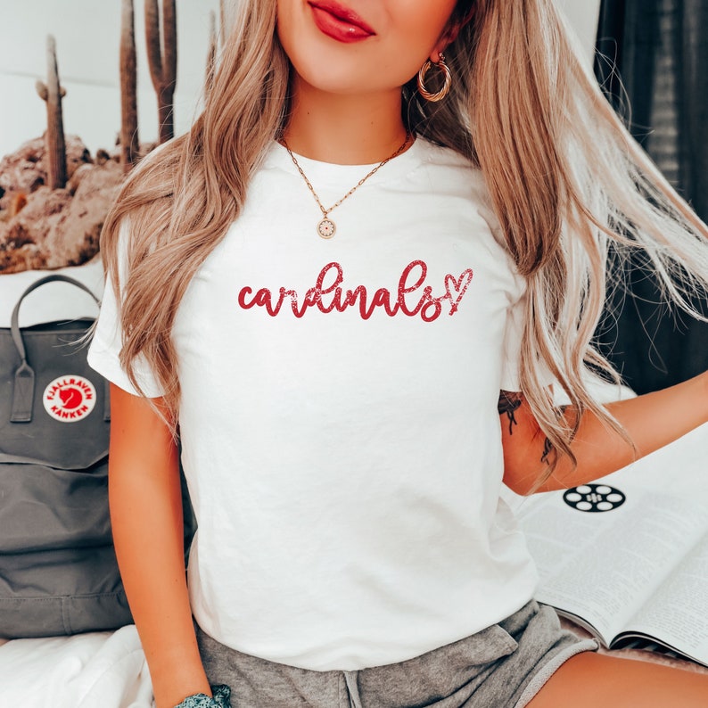 May include: White t-shirt with the text "cardinals" in red script with a red heart next to it.