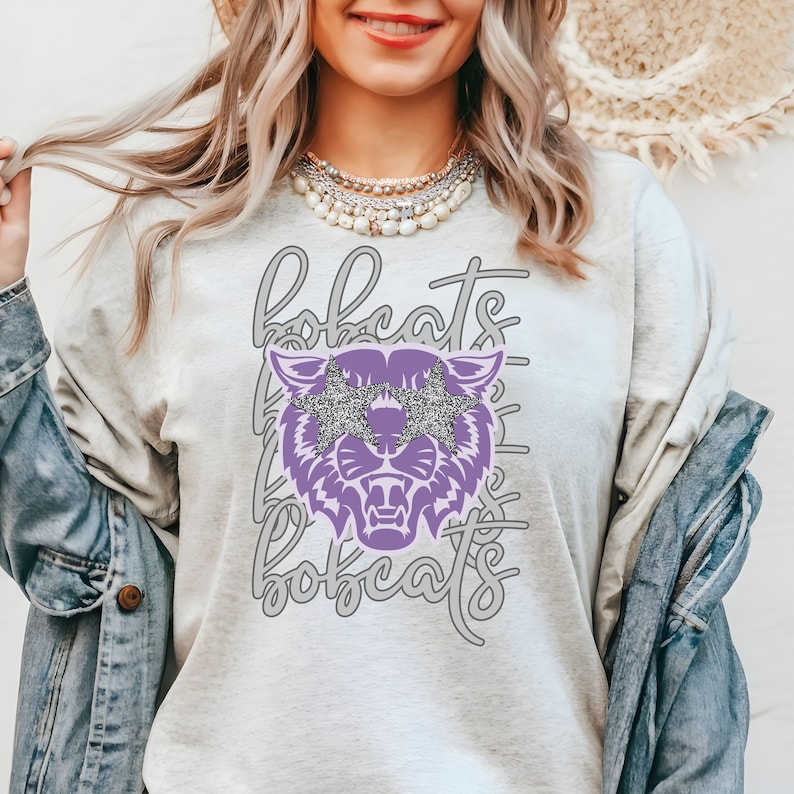 May include: A light gray sweatshirt with the word "bobcats" in a script font. A purple bobcat graphic with glitter star-shaped sunglasses is centered on the front. The word "bobcats" is repeated in a vertical pattern behind the bobcat.