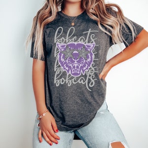 May include: Dark gray t-shirt featuring a purple bobcat graphic with star-shaped glitter sunglasses. The word "bobcats" is written above and below the graphic in a cursive font. The shirt is a crew neck.