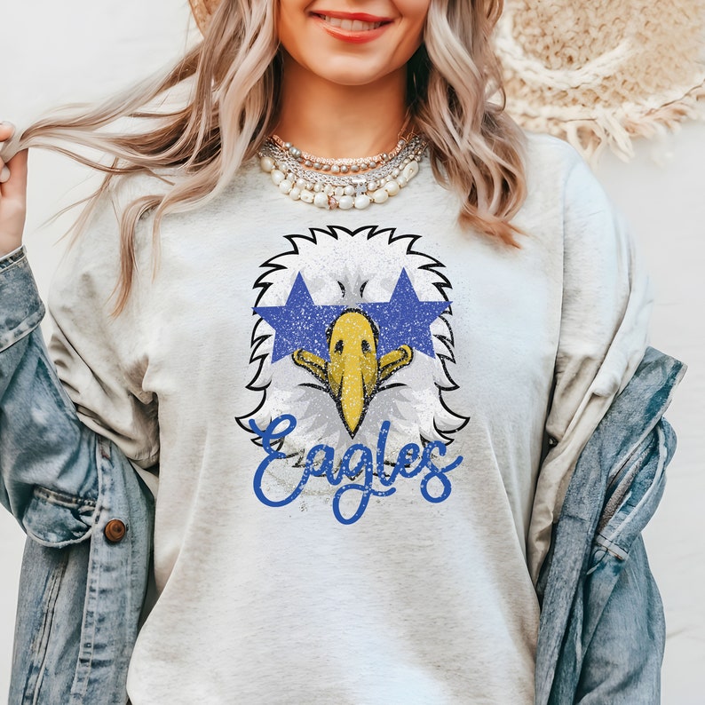 Pu&ograve; includere: Una maglietta grigia con una grafica usurata di un'aquila che indossa occhiali da sole blu a forma di stella e la parola "Eagles" in scrittura blu.