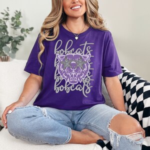May include: A purple t-shirt featuring a stylized bobcat head wearing glitter star-shaped sunglasses. The word "bobcats" is written in a script font above and below the graphic. The shirt is a solid purple color.