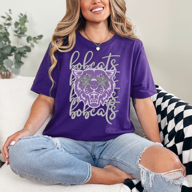 May include: A purple t-shirt featuring a stylized bobcat head wearing glitter star-shaped sunglasses. The word "bobcats" is written in a script font above and below the graphic. The shirt is a solid purple color.