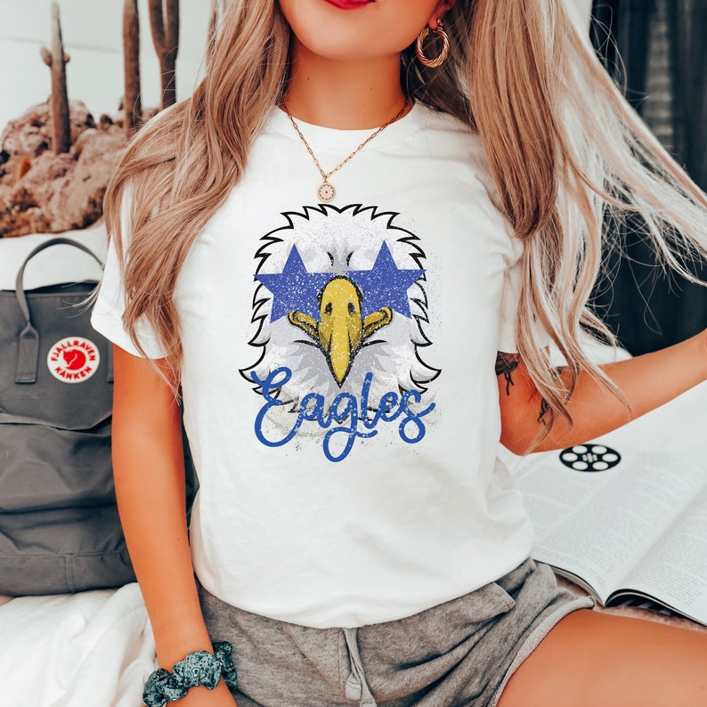 Pu&ograve; includere: Maglietta bianca con una grafica di un'aquila blu e gialla e la parola "Eagles" in lettere blu.