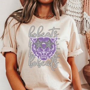 May include: Cream-colored t-shirt featuring a purple bobcat graphic wearing star-shaped sunglasses with silver glitter. The word "bobcats" is repeated in a gray, cursive font. The shirt has short sleeves and a relaxed fit.