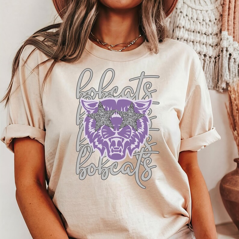 May include: Cream-colored t-shirt featuring a purple bobcat graphic wearing star-shaped sunglasses with silver glitter. The word "bobcats" is repeated in a gray, cursive font. The shirt has short sleeves and a relaxed fit.