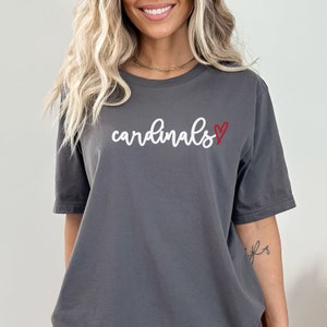 May include: A gray t-shirt with the word "cardinals" written in white cursive with a red heart next to it.