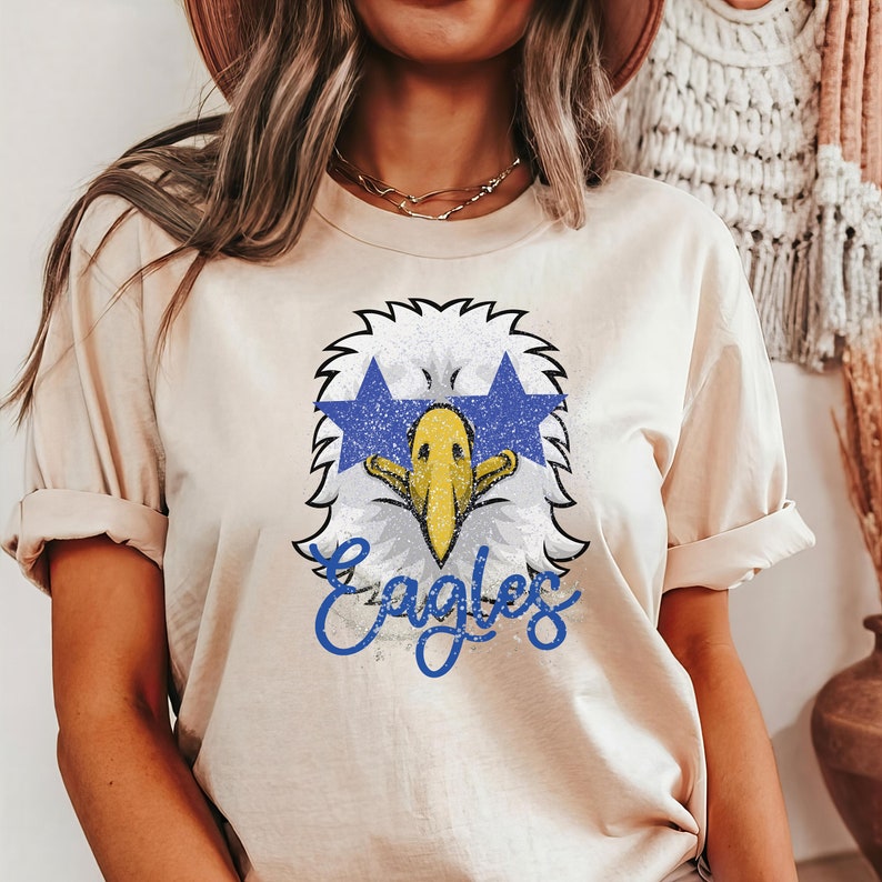 Pu&ograve; includere: Una maglietta beige con una grafica usurata di un'aquila che indossa occhiali da sole blu a forma di stella e la parola "Eagles" in scrittura blu.