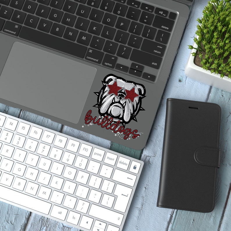 K&ouml;nnte beinhalten: Ein silberner Laptop mit einer wei&szlig;en Tastatur und einem Aufkleber mit einem Bulldoggen, der eine rote, sternf&ouml;rmige Sonnenbrille tr&auml;gt. Der Aufkleber lautet "bulldogs".