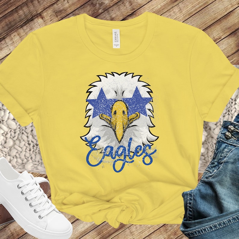 Pu&ograve; includere: Maglietta gialla con una grafica di un'aquila blu e bianca e la parola "Eagles" in lettere blu.