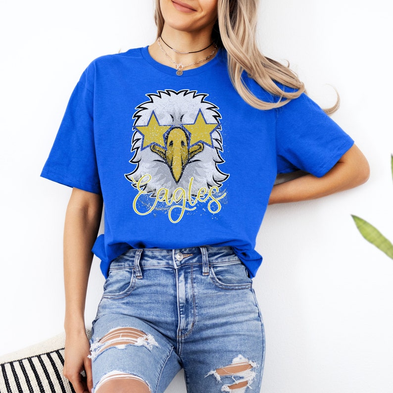 Pu&ograve; includere: Una t-shirt blu reale con una grafica usurata di un'aquila che indossa occhiali da sole e la parola "Eagles" in lettere dorate.