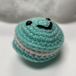 Puede incluir: Un peluche redondo, tejido a ganchillo, de color turquesa, con un centro blanco y ojos y boca bordados en negro. El peluche tiene una cara sonriente. Es un juguete blando.