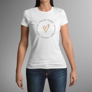 May include: White t-shirt with a circular design. The design includes a gold heart and the words "care giver, change maker, encourager, teacher." The shirt is a crew neck style and is worn with blue jeans.