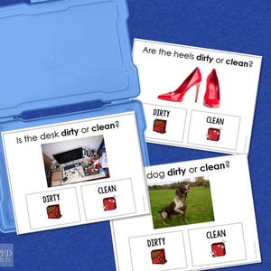 Könnte beinhalten: Eine blaue Kunststoff-Aufbewahrungsbox enthält drei Lernkarten. Die Karten stellen Fragen: „Ist der Schreibtisch schmutzig oder sauber?“ „Sind die Absätze schmutzig oder sauber?“ und „Hund schmutzig oder sauber?“ Jede Karte hat ein Bild und die Optionen „DRECKIG“ und „SAUBER“.