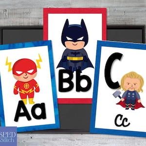 May include: Three framed superhero-themed alphabet art prints. The Flash, Batman, and Thor are depicted with their corresponding letters. The Flash is on a blue frame, Batman on a red frame, and Thor on a blue frame. The letters are in a bold, black font.