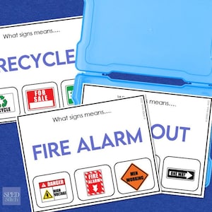 Peut inclure: Cartes éducatives avec des panneaux de signalisation et de sécurité, dont "RECYCLE", "FIRE ALARM" et "ONE WAY". Les cartes sont blanches avec du texte bleu et des illustrations colorées, rangées dans une boîte en plastique bleu clair.
